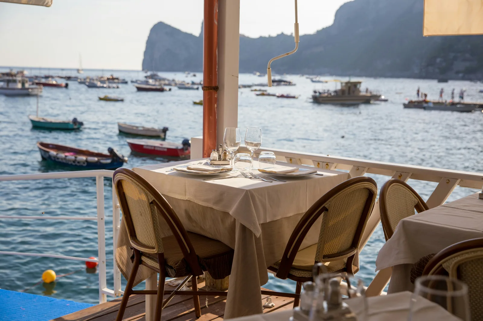 A linen-laid restaurant table on a balcony overlooking a marina of small wooden boats in Capri.