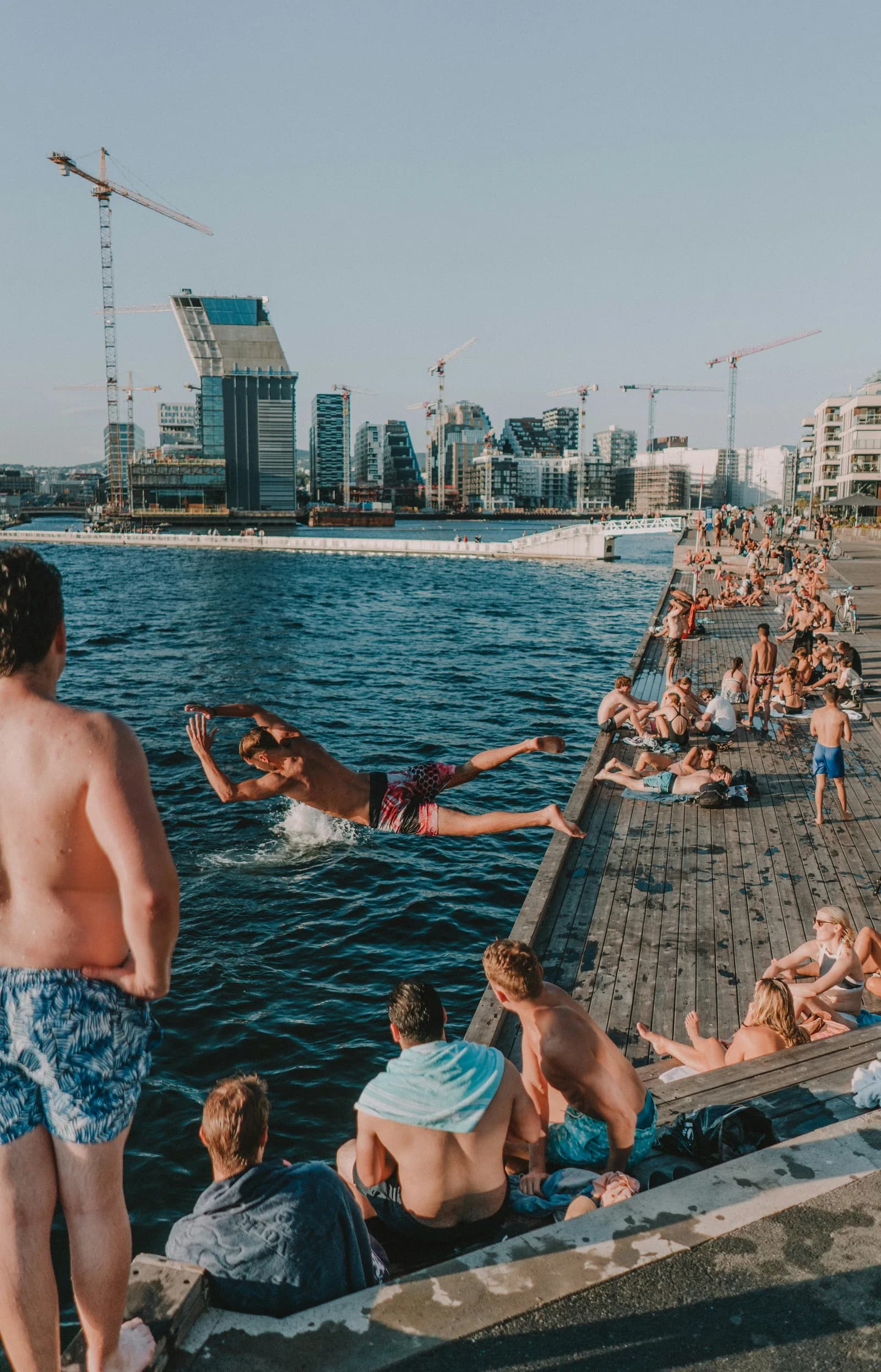 Swimmers diving into the Oslofjord from the wooden Sørenga harbour-bath deck, the Bjørvika skyline beyond on a summer evening.